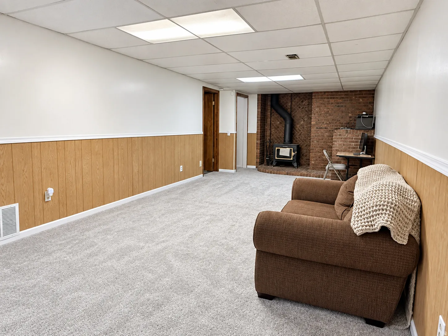 Finished room featuring wood-finish paneling, light-colored carpeting, a drop ceiling with recessed lighting, and a brick accent wall with a wood-burning stove
