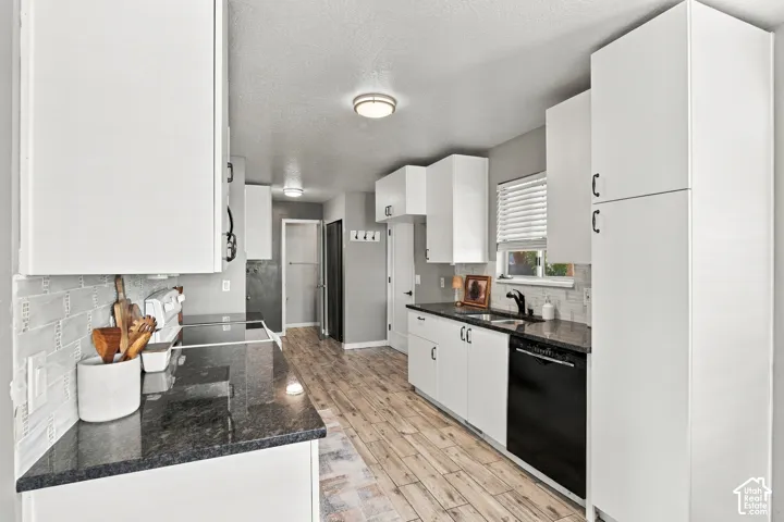 Kitchen featuring white cabinetry, dark countertops, wood-finish flooring, a tile backsplash, and a built-in dishwasher