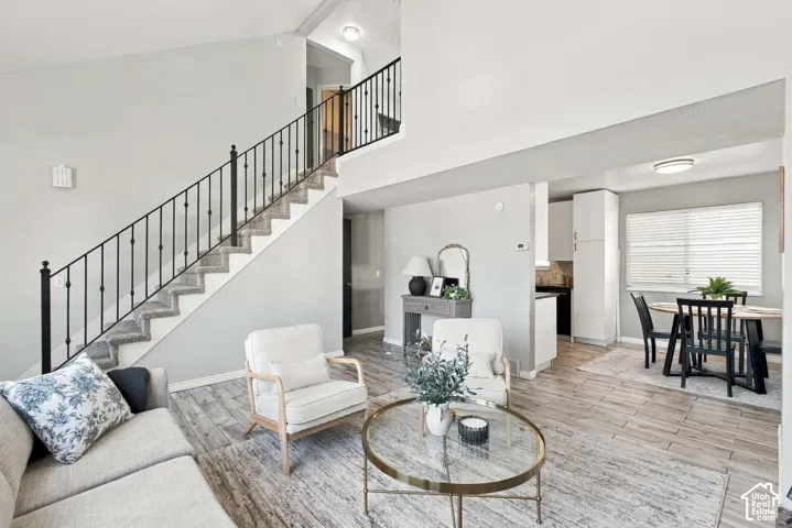 Expansive living area with wood-finish flooring, a carpeted staircase with iron railings, and a dining area featuring a window with blinds