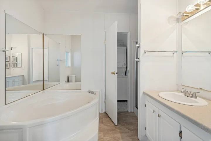Corner soaking tub with surrounding wall mirrors, vanity with an integrated sink, built-in cabinetry, and a stacked washer and dryer unit