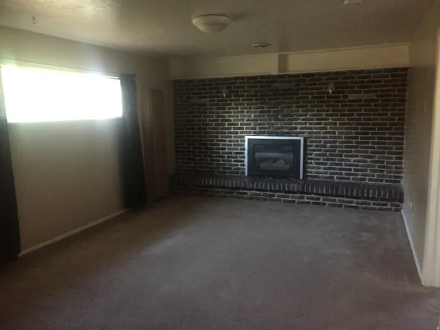 Carpeted room featuring a brick accent wall with a built-in fireplace and hearth, illuminated by a window. Photo shown was taken prior to tenant occupancy and is provided for reference.