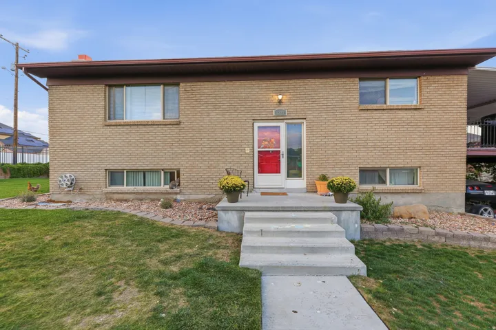 Brick exterior with a red entry door, concrete front steps, and a landscaped front yard featuring a grass lawn, larger bricks and stone  filled flower bed