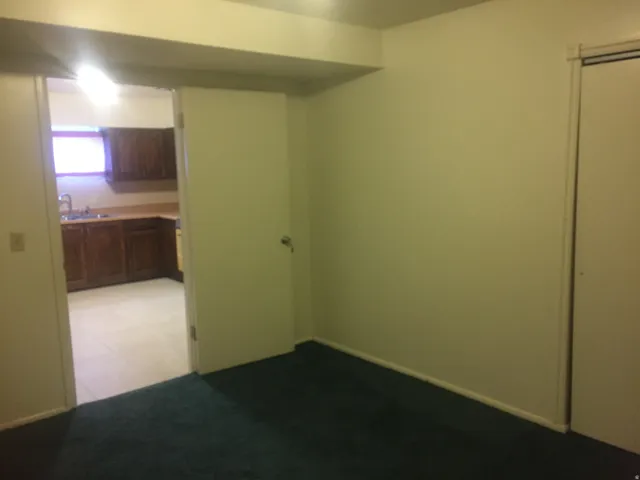 Dark carpet flooring, white walls, and a white door with a silver handle, view from east side basement bedroom into second kitchen. Photo shown was taken prior to tenant occupancy and is provided for reference.