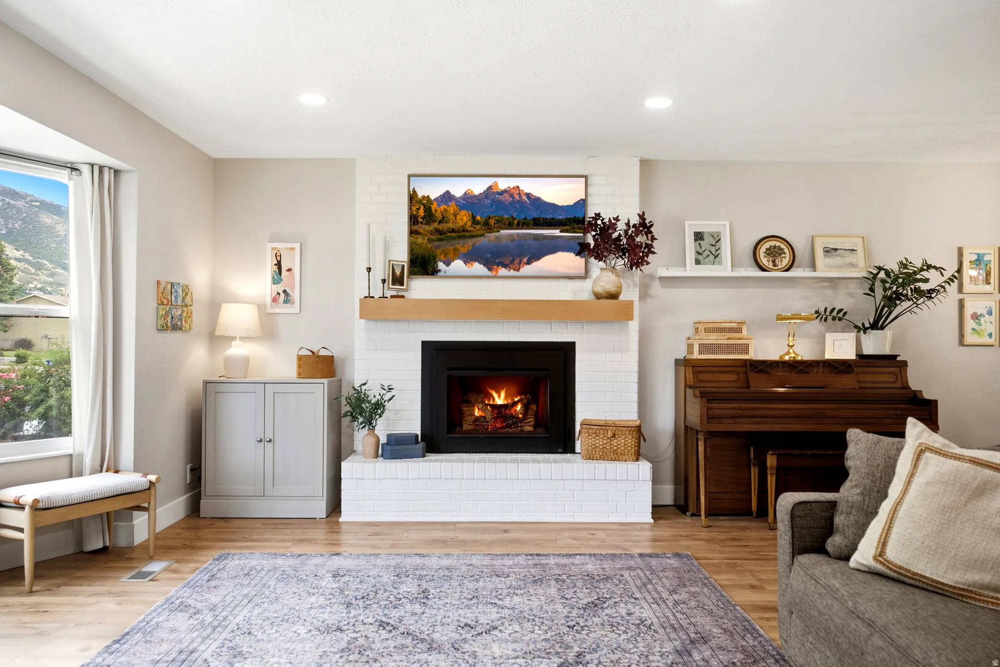 Recessed lighting, wood-finish flooring, white brick fireplace with a wood mantle, large window with natural light, and a built-in floating shelf