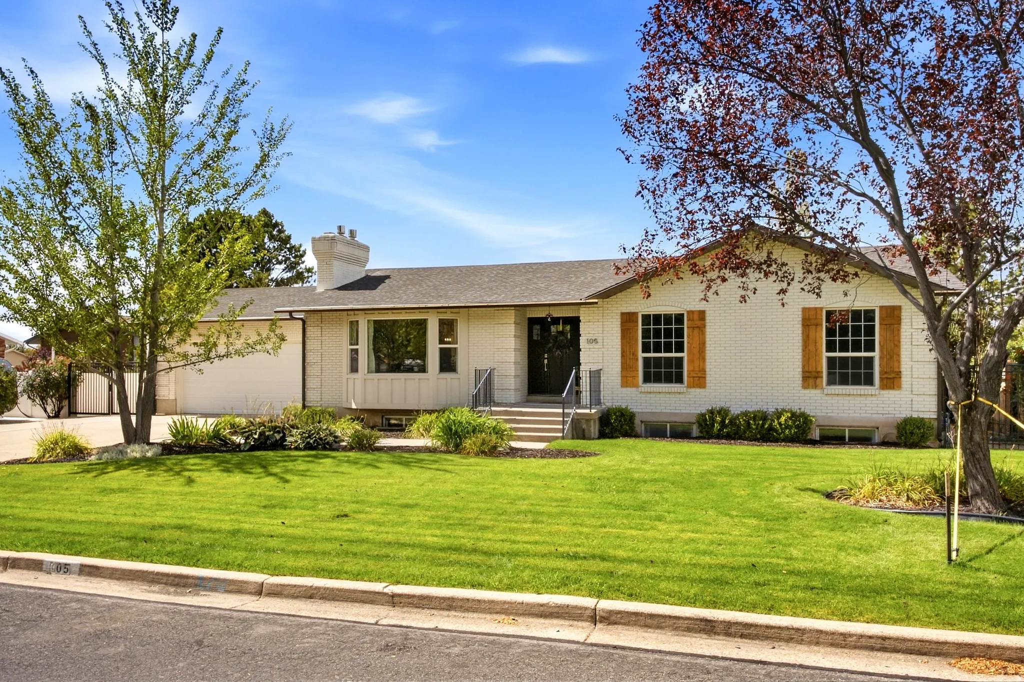 White brick exterior with wood shutters, front entry with steps and wrought iron railing, attached garage, and landscaped front yard with mature trees