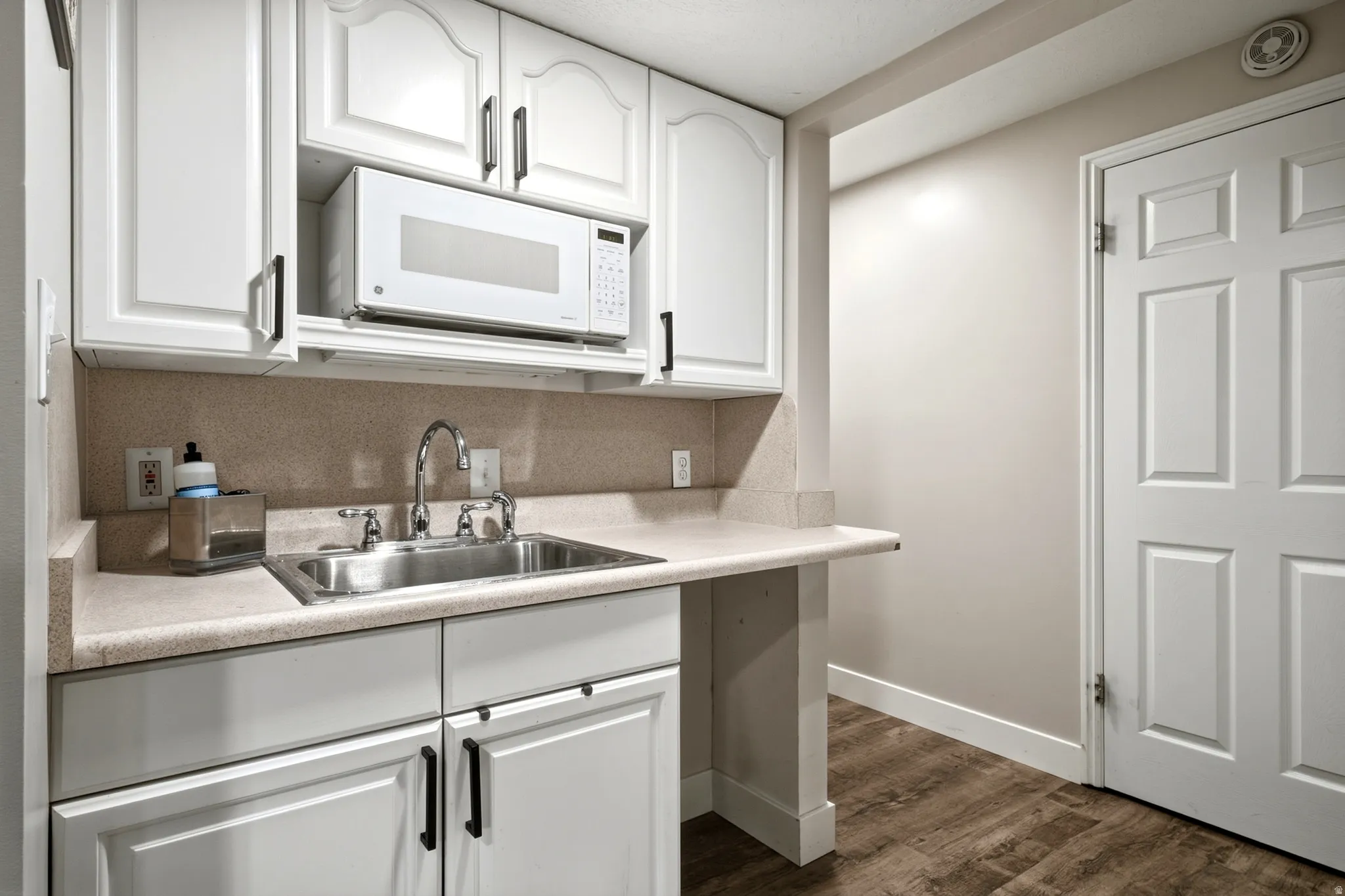 White cabinetry with black hardware, stainless steel sink with chrome faucet, built-in microwave, wood-finish flooring, and a six-panel interior door