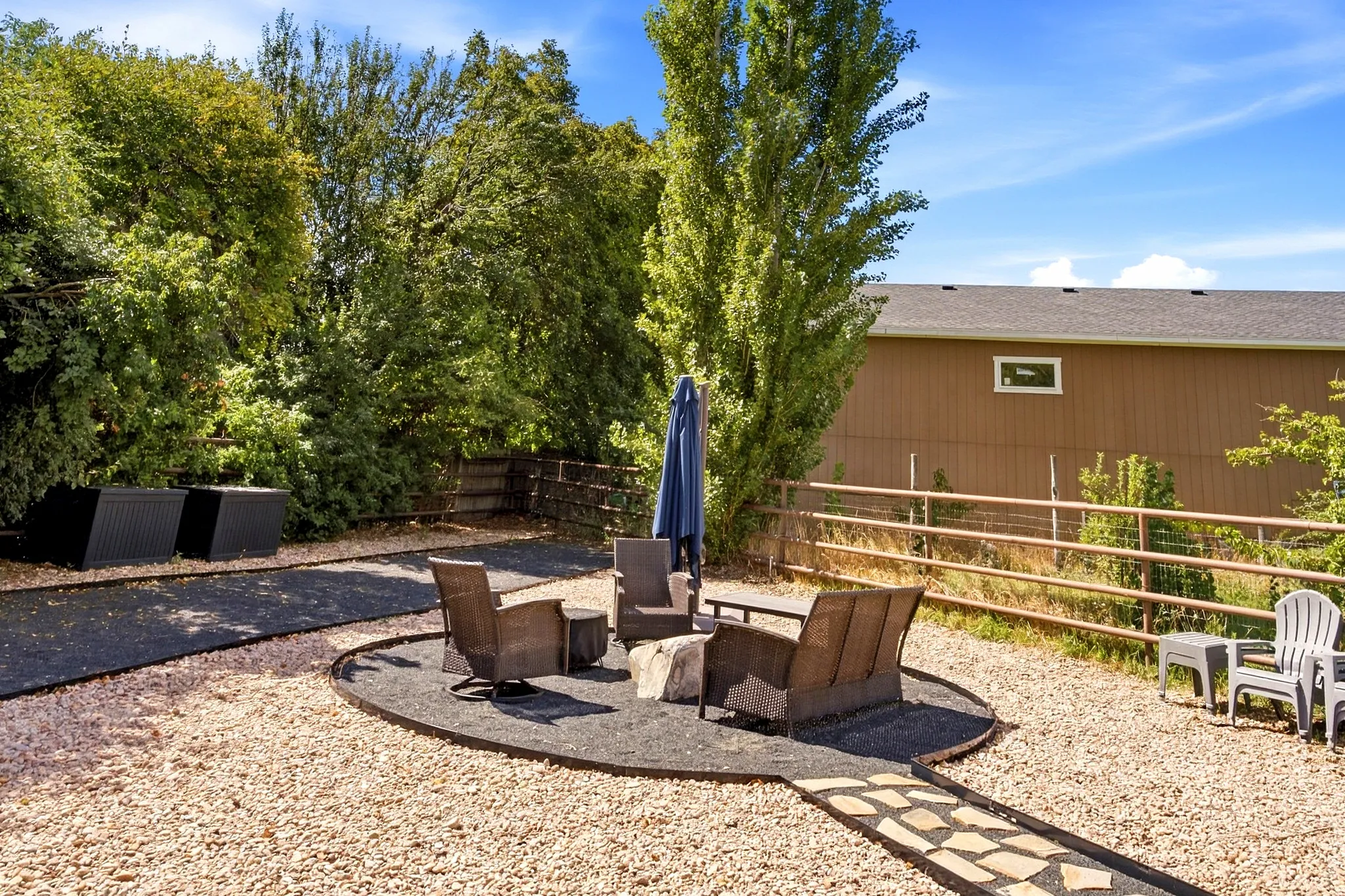 Expansive outdoor space featuring a large gravel area, a defined fire pit zone with dark ground cover, a stone pathway, and mature trees providing natural screening
