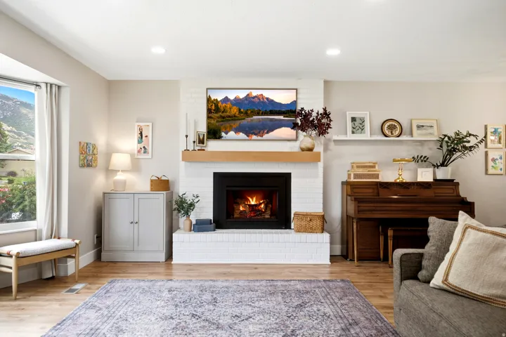 Recessed lighting, wood-finish flooring, white brick fireplace with a wood mantle, large window with natural light, and a built-in floating shelf