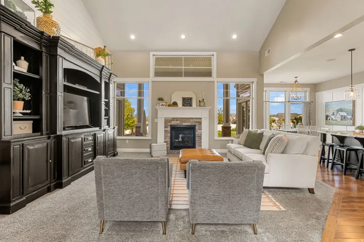 Vaulted ceilings with recessed lighting, stone-surround fireplace with mantel, large picture windows, built-in entertainment cabinetry, and wood-finish flooring