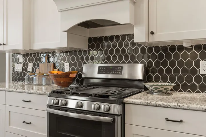 Stainless steel range with a built-in griddle, granite-style countertops, white shaker-style cabinetry, dark hexagonal tile backsplash, and a custom range hood
