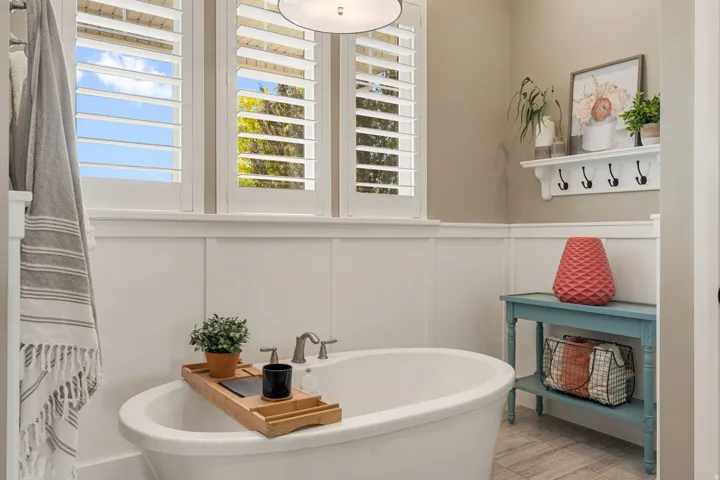 Freestanding bathtub with deck-mounted faucet, plantation shutters, board and batten wainscoting, and wood-finish flooring
