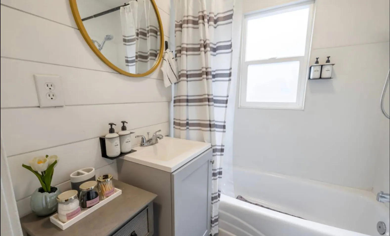 White shiplap wall paneling, gold-framed round mirror, integrated sink vanity, white bathtub, and frosted window