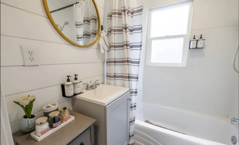White shiplap wall paneling, gold-framed round mirror, integrated sink vanity, white bathtub, and frosted window