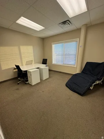 Unit D- Windowed room with textured carpet flooring, drop ceiling with integrated lighting, and a neutral wall palette