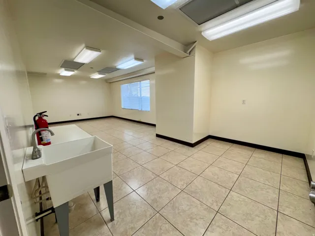 Unit A- Spacious room with light-toned tiled flooring, multiple overhead fluorescent light fixtures, and a utility sink with a faucet