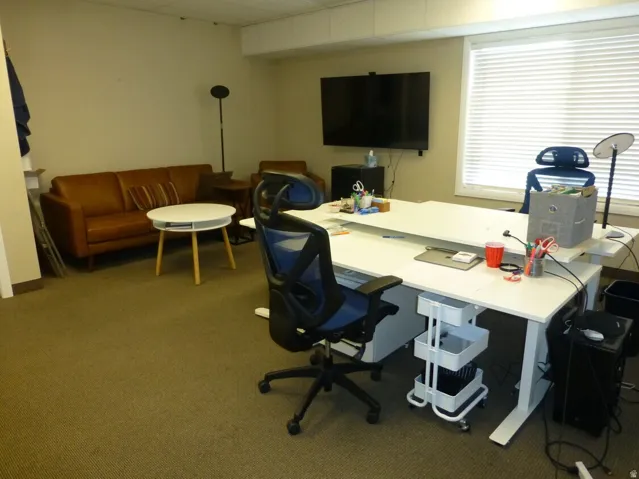 Unit D- Wall-mounted display, window with horizontal blinds, and carpeted flooring
