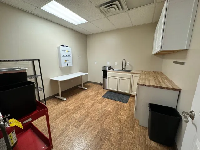 Wood-finish flooring, drop ceiling with rectangular overhead light, wood-finish countertops, stainless steel sink with gooseneck faucet, and white cabinetry
