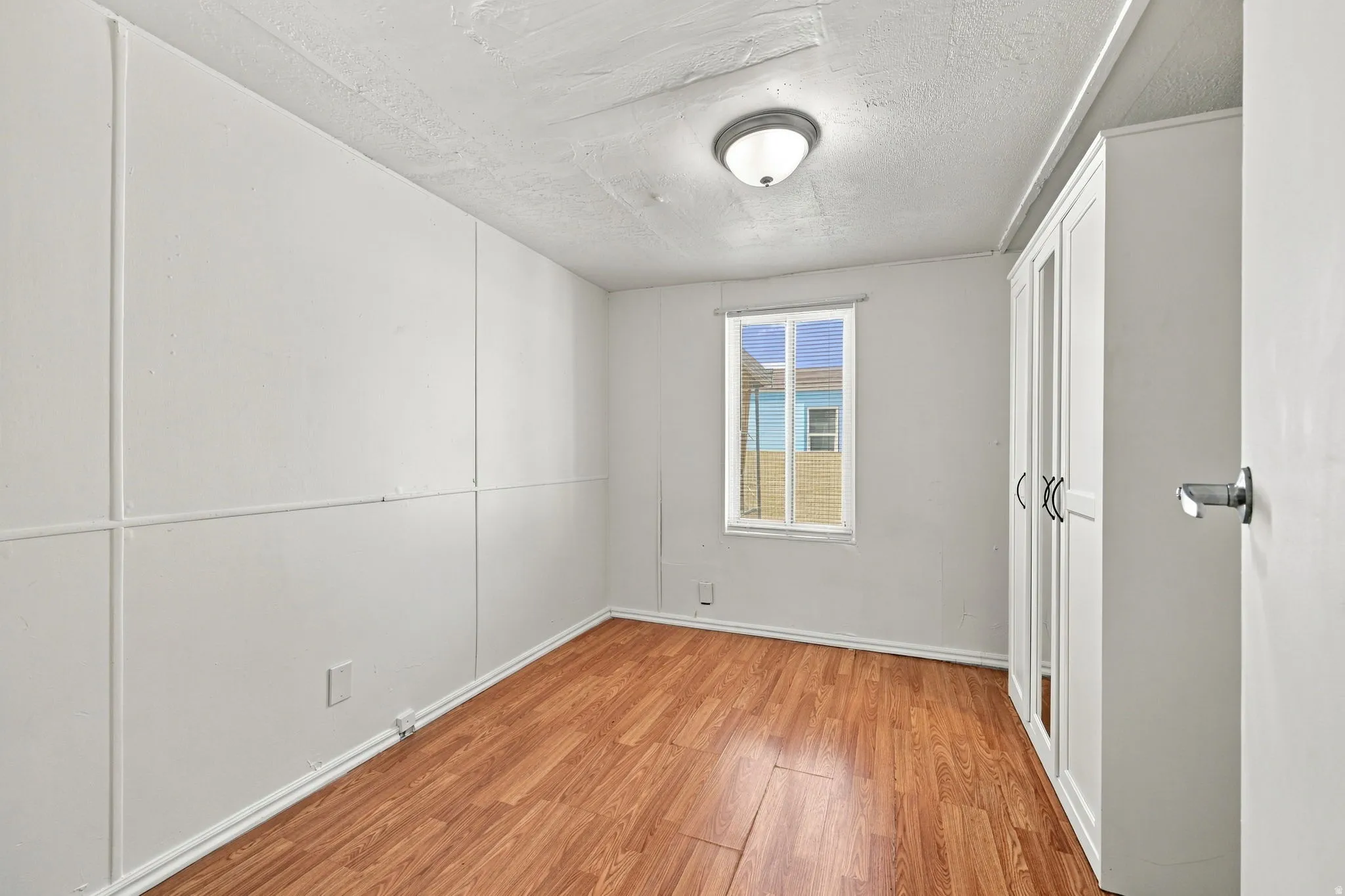 Wood-finish flooring, white paneled walls, ceiling-mounted light fixture, built-in storage cabinet with mirrored doors, and a double-paned window