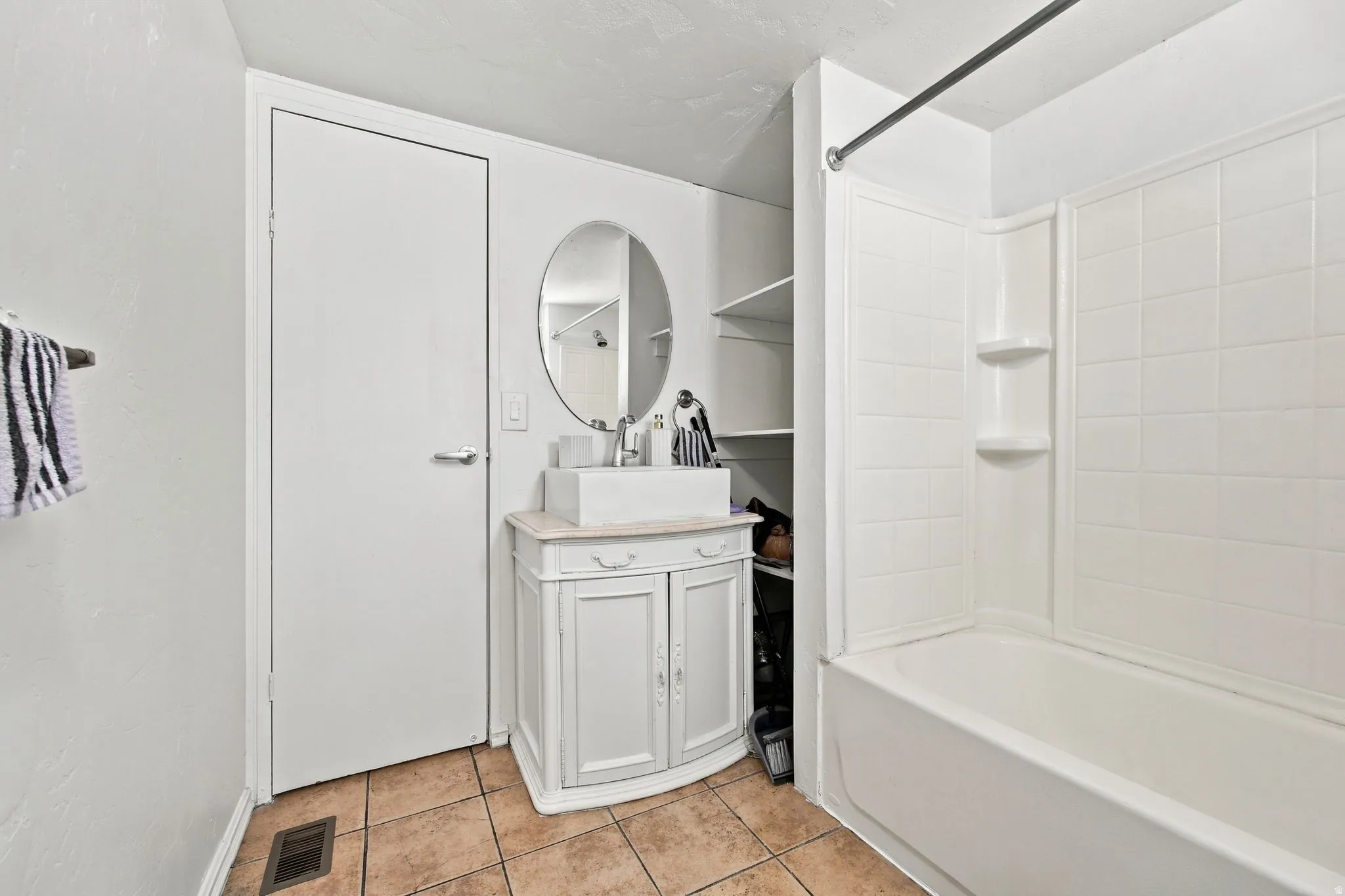 Tile flooring, a white vanity with a vessel sink, an oval mirror, open shelving, and a white tub with a shower surround