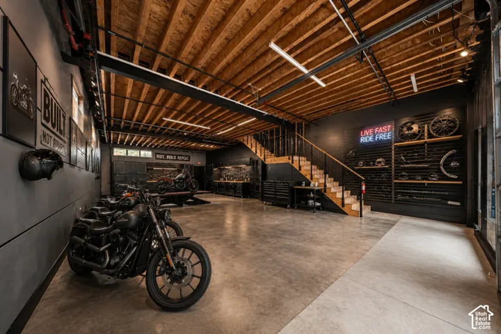 Expansive garage space with exposed wood beam ceiling, concrete flooring, built-in display shelving, and a wood staircase with a metal railing
