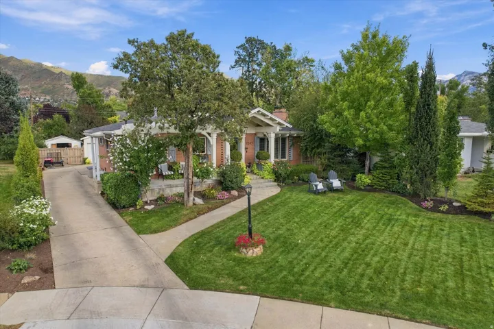 Brick exterior with white trim, a covered porch, a concrete driveway, a manicured lawn, and mature landscaping
