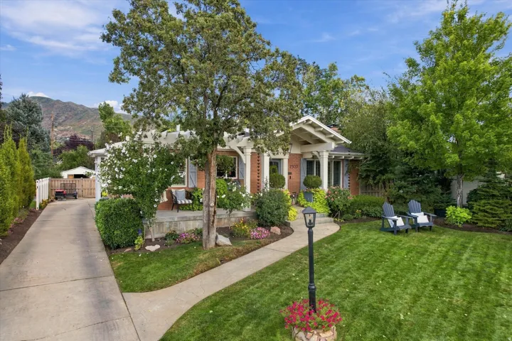 Brick exterior with white pergola accents, mature trees, manicured lawn, concrete driveway, and established landscaping