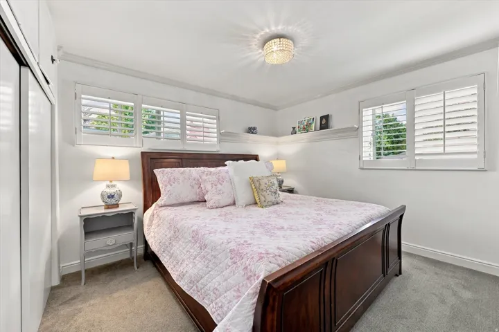 Recessed windows with plantation shutters, crown molding, built-in shelving, wood-finish bed frame, and a flush mount chandelier