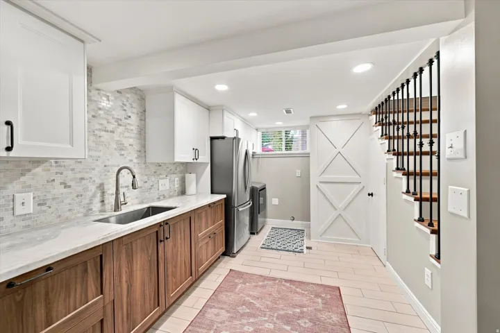 Two-tone cabinetry with white upper cabinets and wood-finish lower cabinets, stainless steel sink with gooseneck faucet, full-size stainless steel refrigerator, white tile backsplash, and light wood-finish flooring