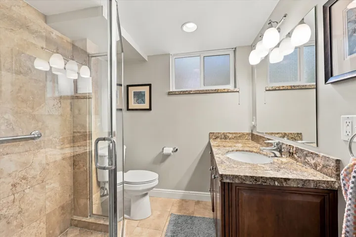 Glass-enclosed shower with stone-look tile surround, vanity with stone-finish countertop, single basin sink, and wood-finish cabinetry
