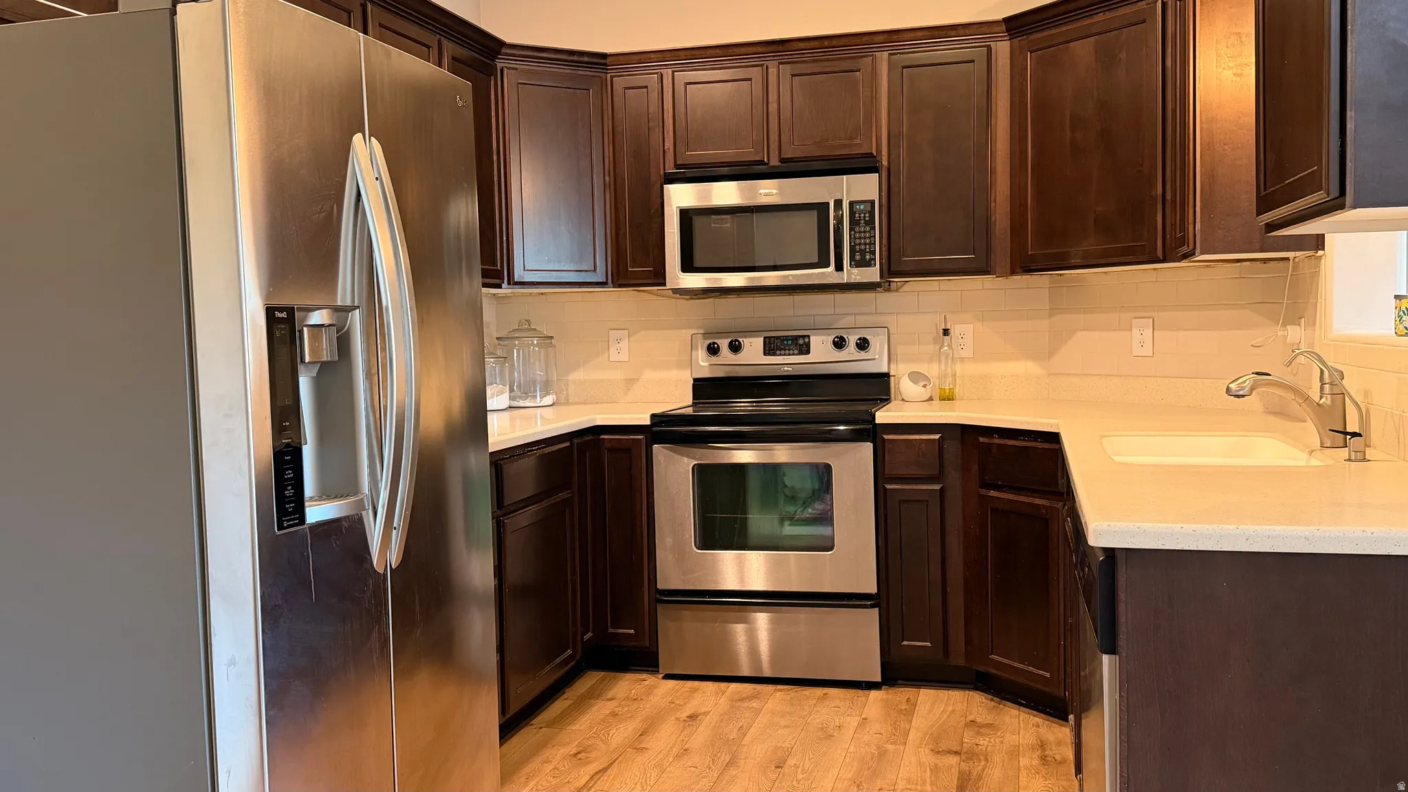 Stainless steel appliances, dark wood cabinetry, light-toned countertops, and wood-finish flooring