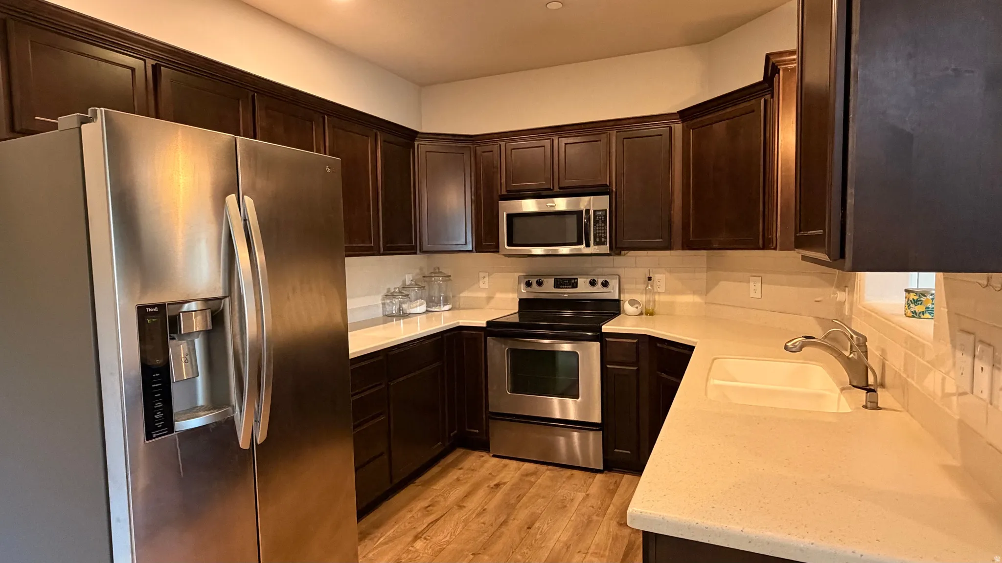 Stainless steel appliances, dark wood-finish cabinetry, light-toned countertops, tile backsplash, and wood-finish flooring