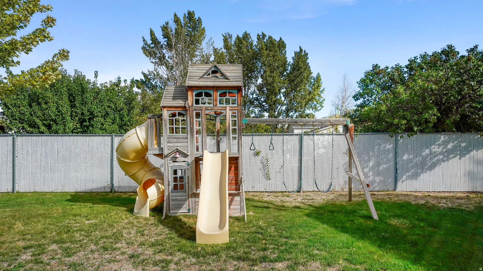 Fenced backyard with a playset featuring a tower, slide, and tube slide, alongside a separate swing set