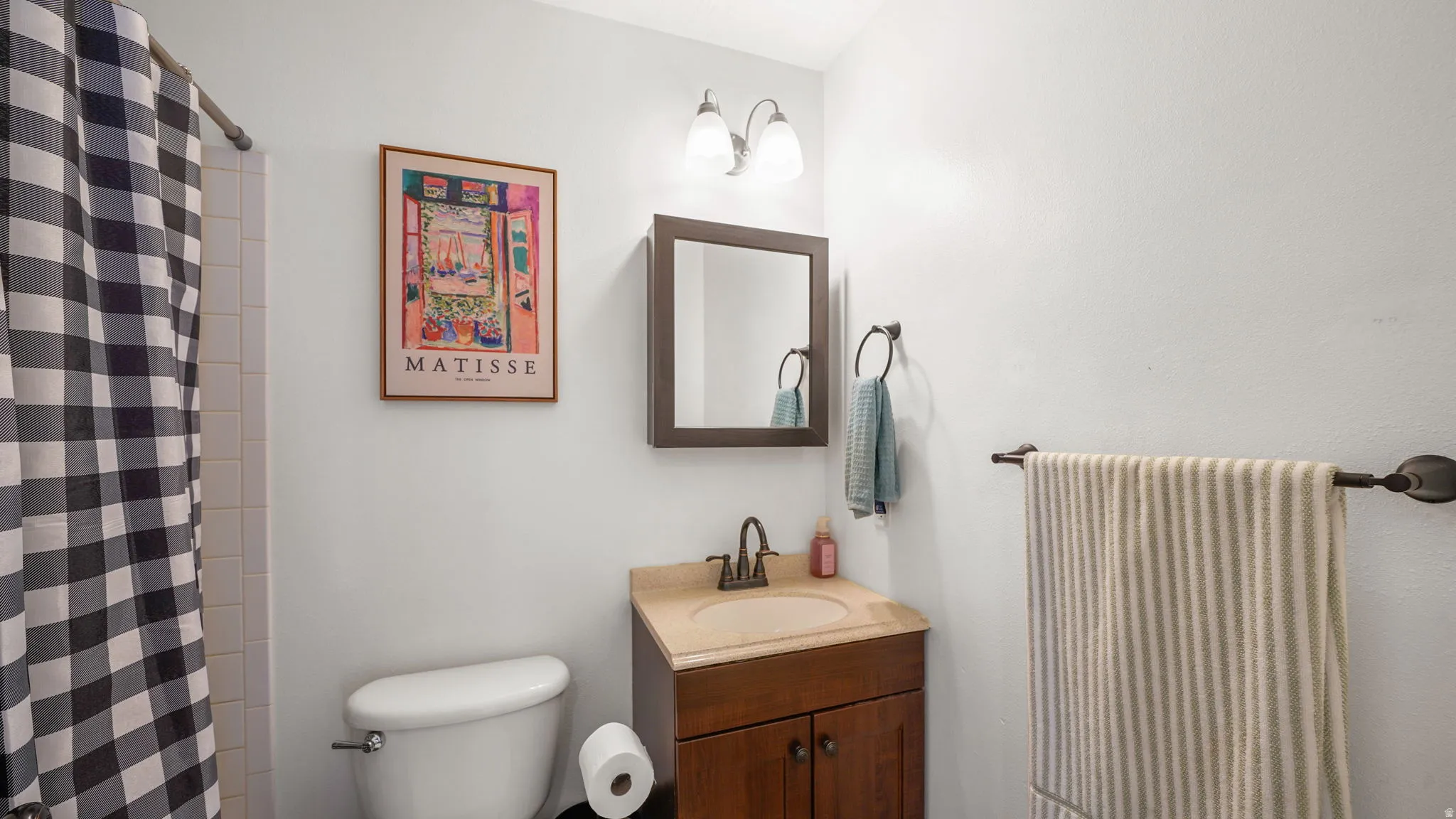 White tile shower surround, single vanity with wood-finish cabinetry, countertop with integrated sink, mounted mirror, and wall-mounted light fixture