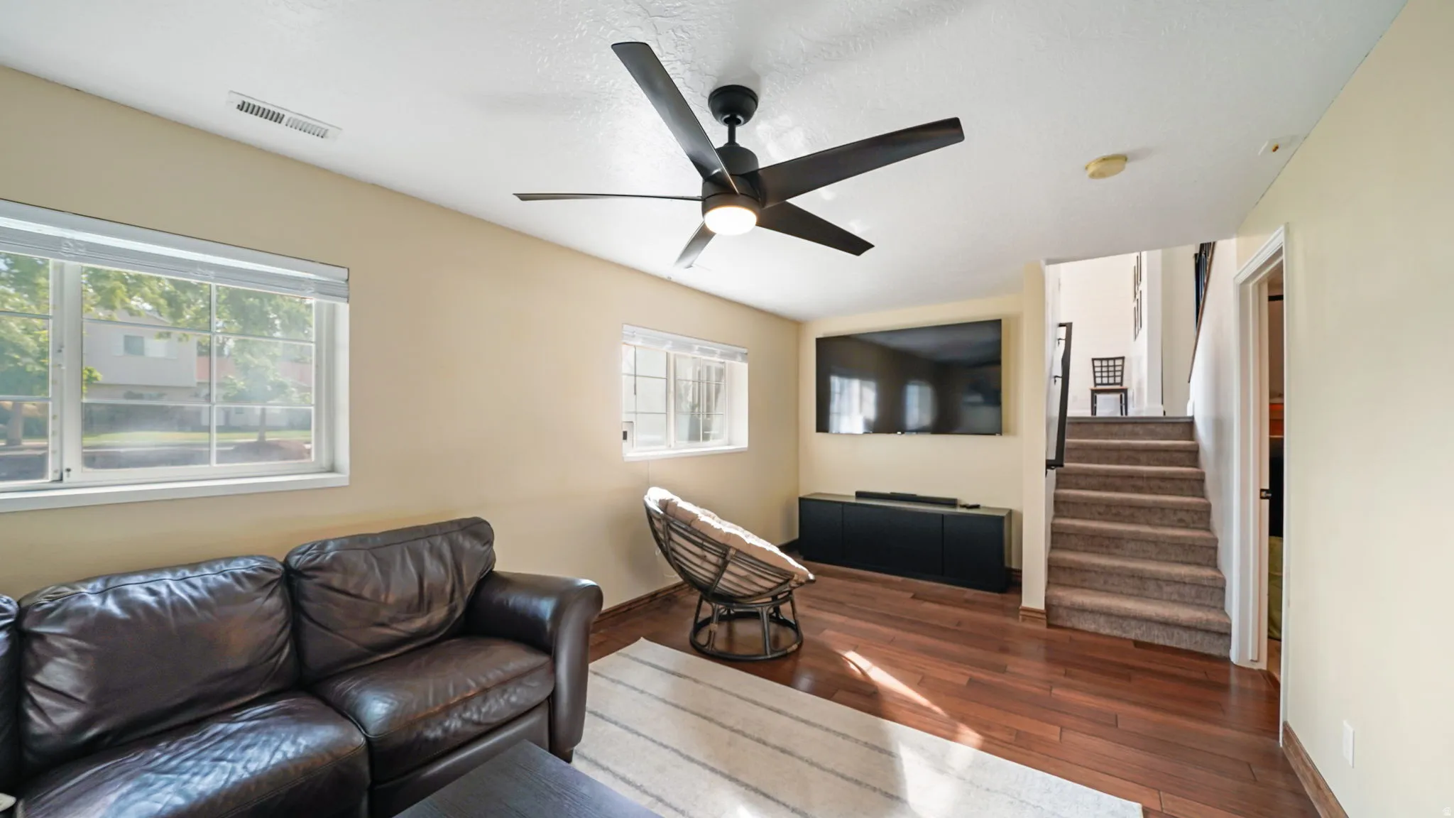 Wood-finish flooring, ceiling fan with integrated lighting, multiple windows, built-in media console, and a carpeted staircase