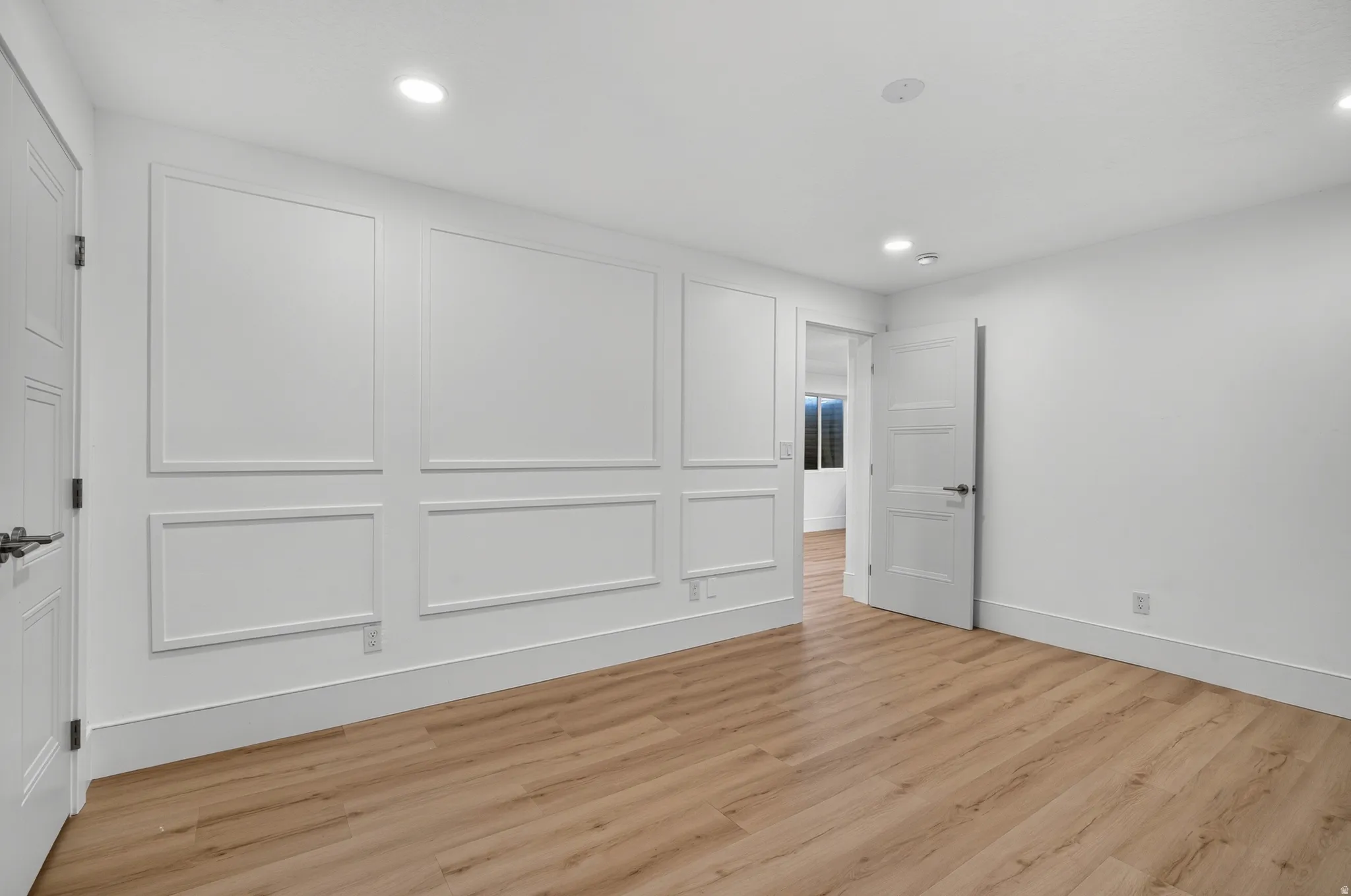 Light wood-finish flooring, recessed lighting, white walls, decorative wall paneling, and a solid-core door with contemporary hardware