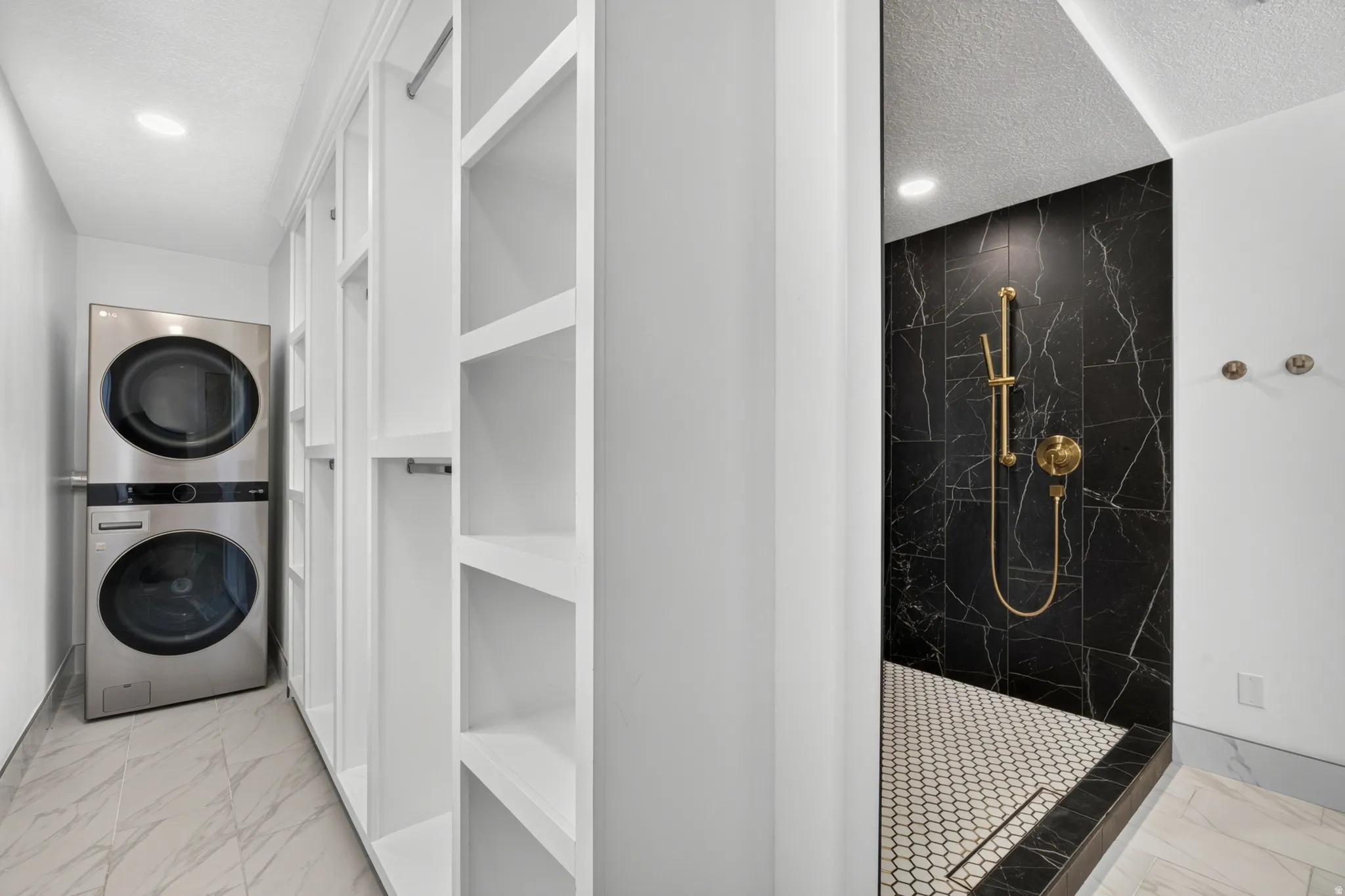 Built-in shelving, stacked laundry appliances, black marble-patterned shower tile, gold-finish shower fixtures, and hexagonal shower floor tile