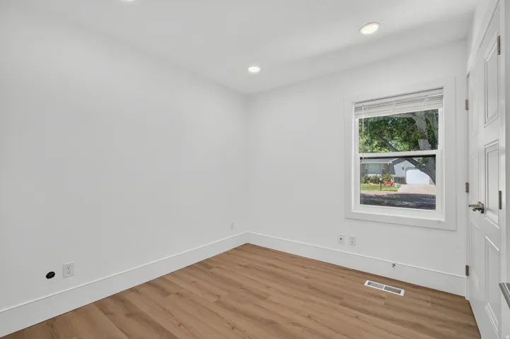 Recessed ceiling lighting, wood-finish flooring, single window with blinds, panel door, and white painted walls