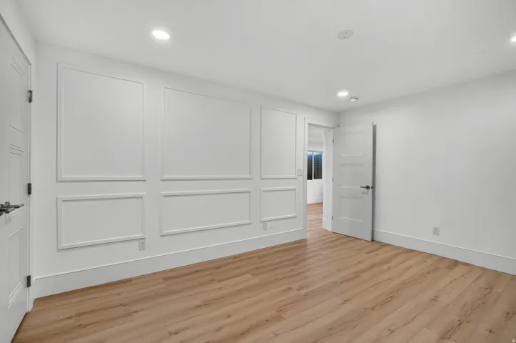Light wood-finish flooring, recessed lighting, white walls, decorative wall paneling, and a solid-core door with contemporary hardware