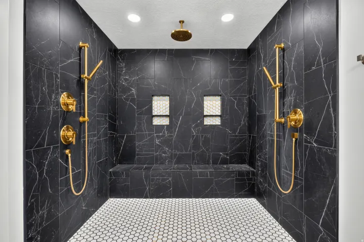 Shower with black marble-look tile walls, dual shower heads, a built-in bench, recessed niches, and white hexagonal floor tiles