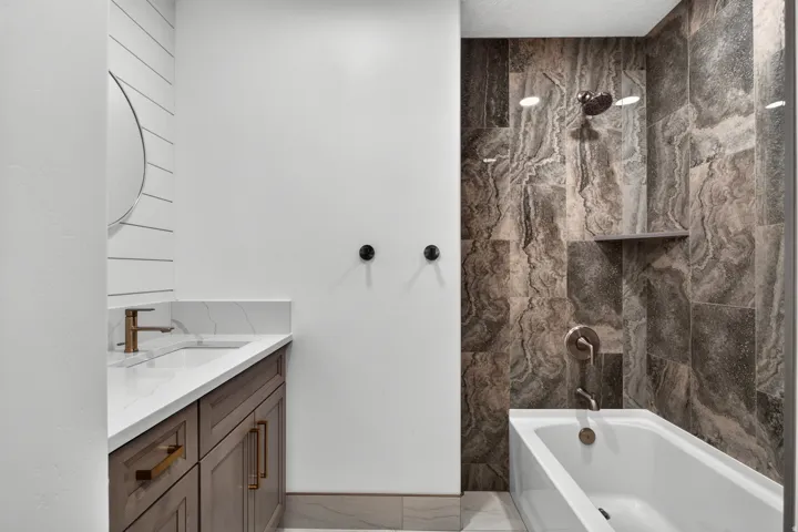 Wood-finish vanity with gold-tone hardware, white countertop with integral sink, ship-lap accent wall, and a tiled shower/tub combination with a built-in shelf