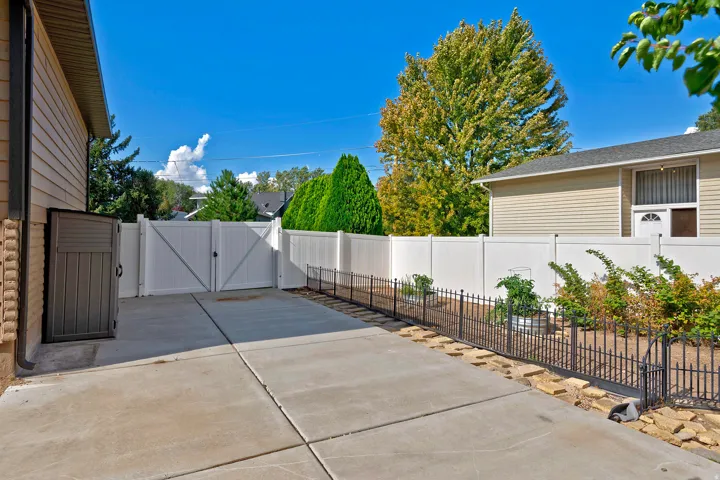 Concrete driveway with a double white gate, flanked by a black metal fence and mature shrubbery