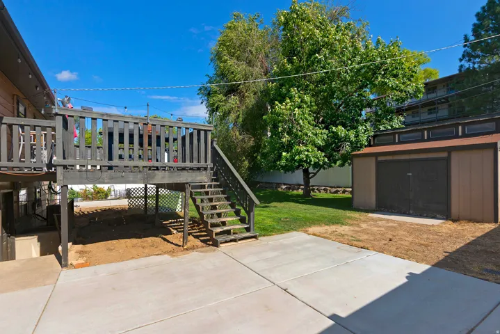 Expansive concrete patio with a raised wood deck and stairs, featuring a storage shed and mature trees