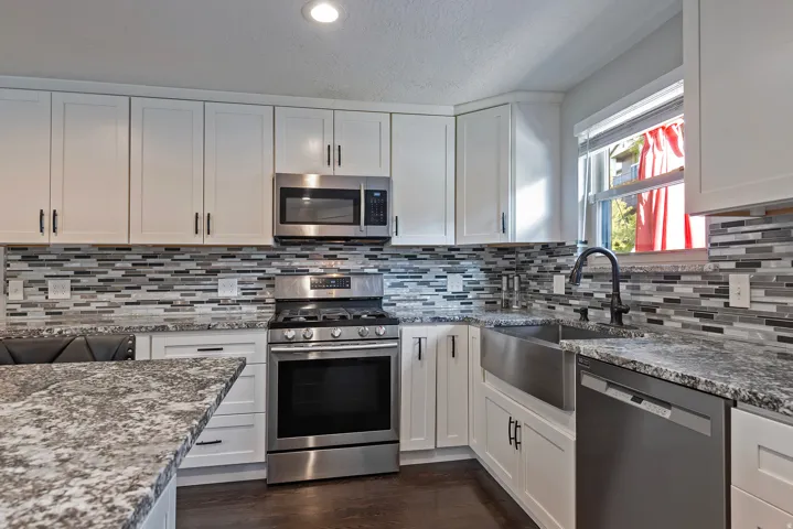 White cabinetry with dark hardware, stainless steel appliances, granite countertops, tile backsplash, and a farmhouse-style sink