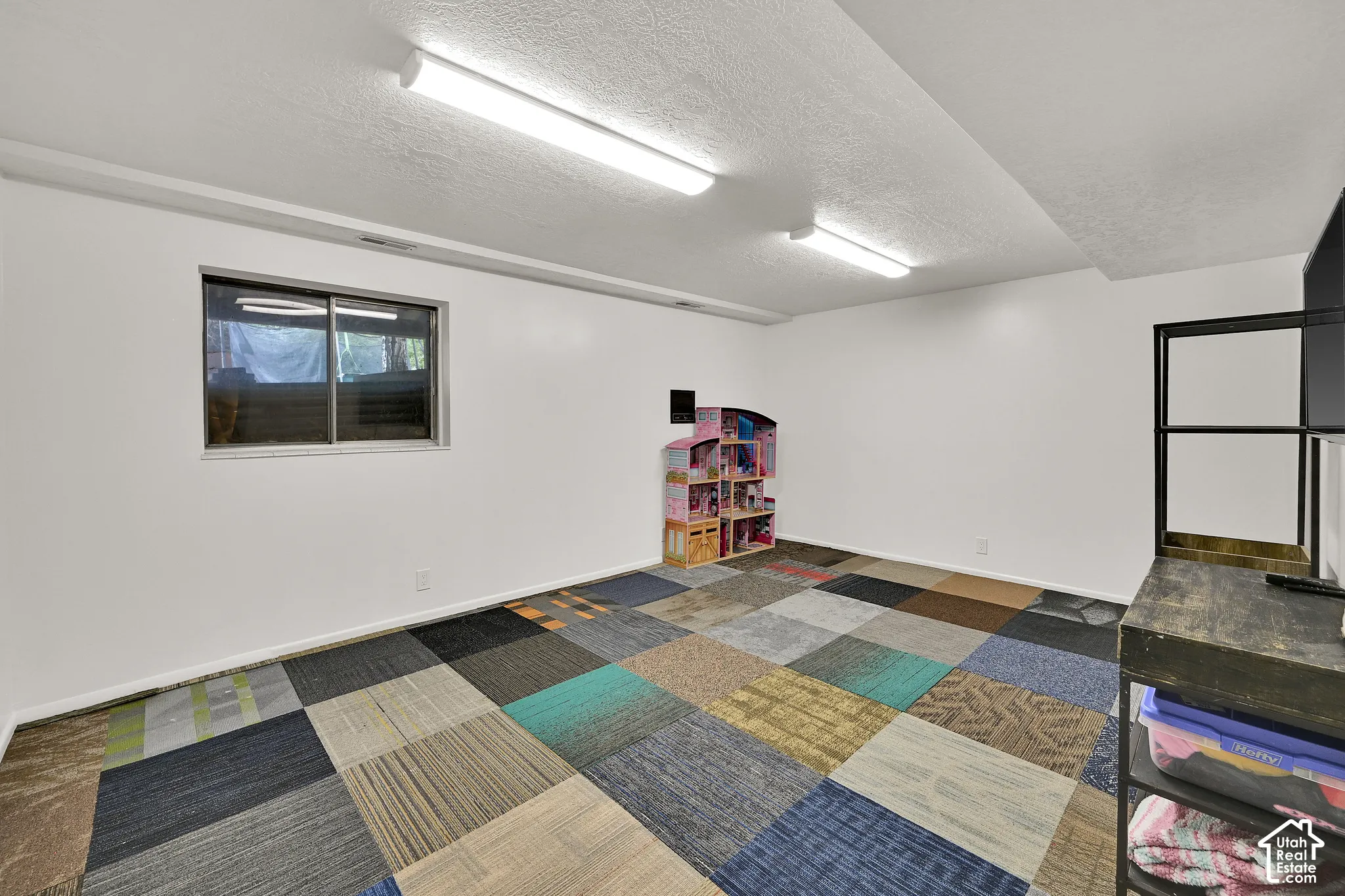 Window with dark frame, white walls, two ceiling-mounted fluorescent light fixtures, and multi-colored square carpet tiles