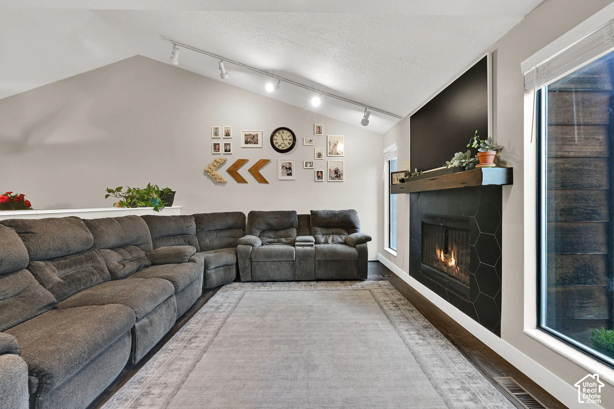 Vaulted ceiling with track lighting, wood-finish flooring, fireplace with hexagonal tile surround, wood mantel, and large window