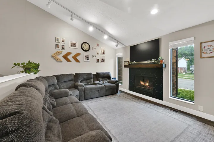 Vaulted ceiling with track lighting, fireplace featuring hexagonal black tile surround, wood mantel, and large windows