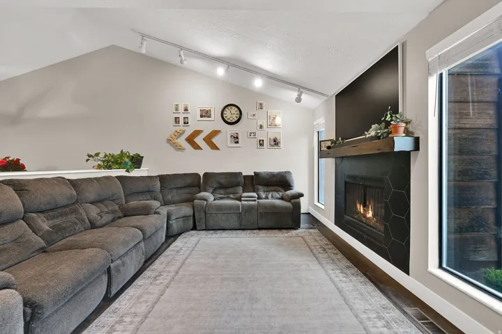 Vaulted ceiling with track lighting, wood-finish flooring, fireplace with hexagonal tile surround, wood mantel, and large window