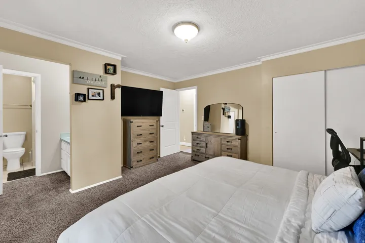 Carpeted room with crown molding, a built-in closet with sliding doors, and access to a bathroom with a white toilet and a vanity with a light-colored countertop