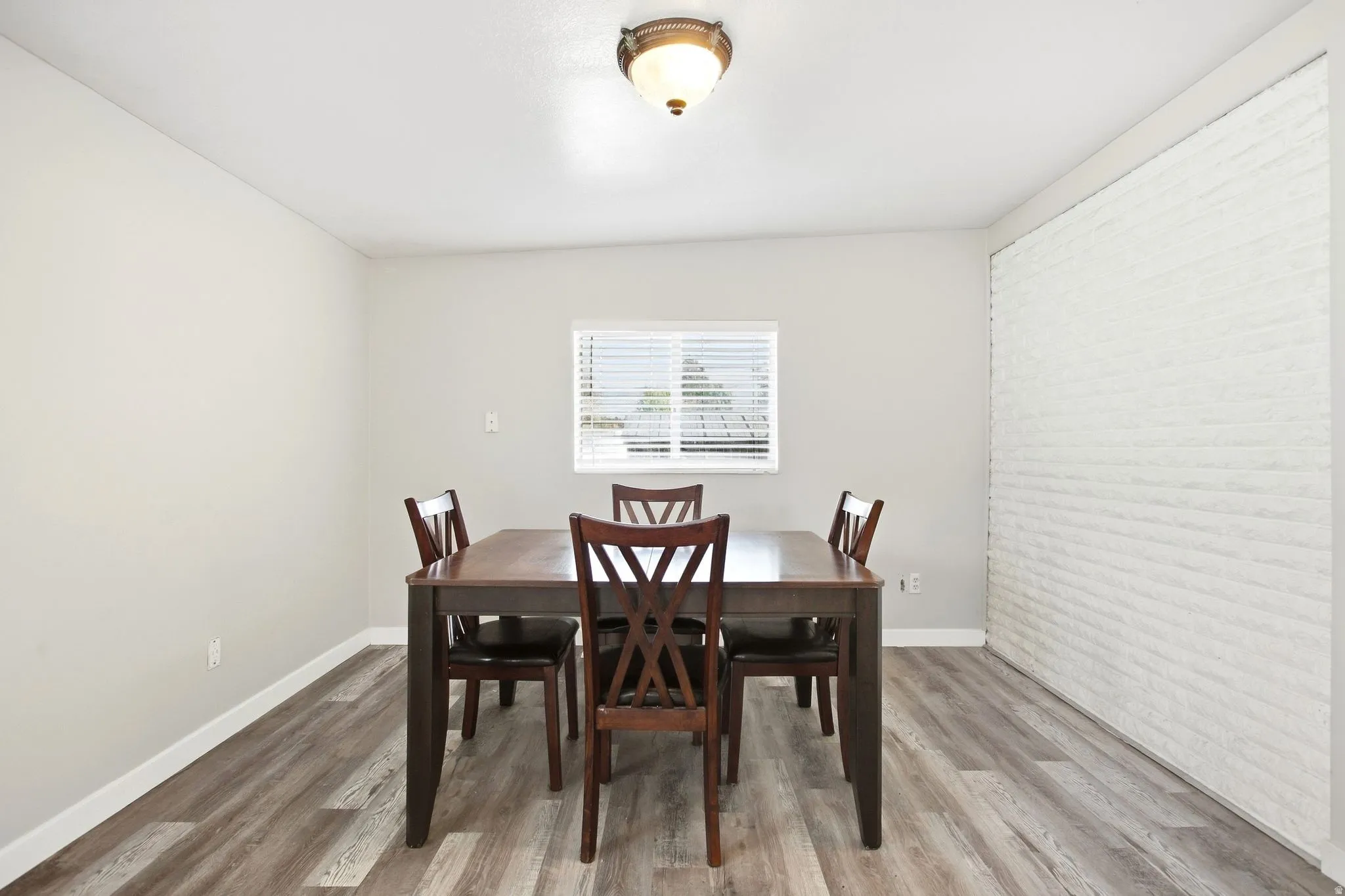 Recessed window with blinds, wood-finish flooring, white brick-pattern accent wall, neutral wall paint, and a ceiling-mounted light fixture