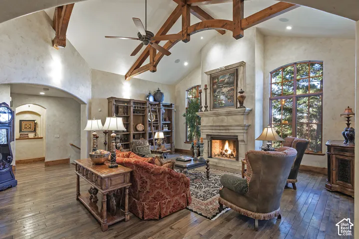 Vaulted ceiling with exposed wood beams, hardwood flooring, stone fireplace with mantel, and arched windows